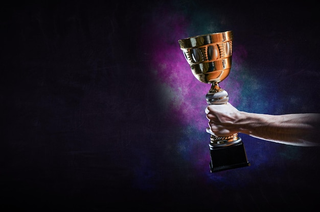 Hand holding up a gold trophy cup against dark background. Mixed media