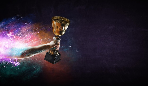 Hand holding up a gold trophy cup against dark background. Mixed media