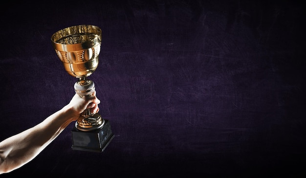 Hand holding up a gold trophy cup against dark background. Mixed media