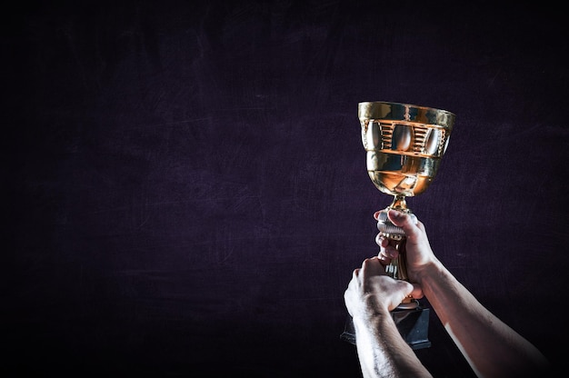 Hand holding up a gold trophy cup against dark background. Mixed media