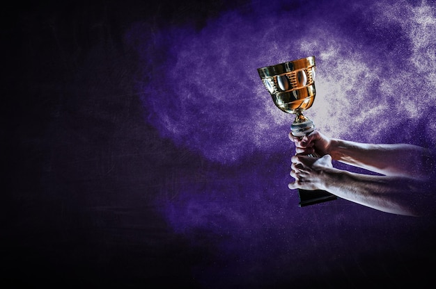 Hand holding up a gold trophy cup against dark background. Mixed media