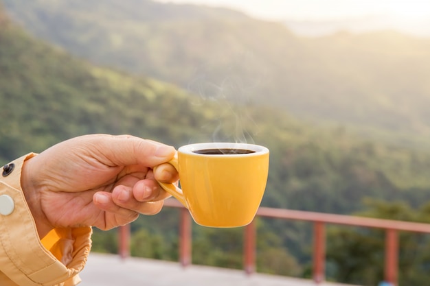 Hand holding a yellow cup of hot espresso coffee mugs and nature view of the mountain landscape in the morning with sunlight