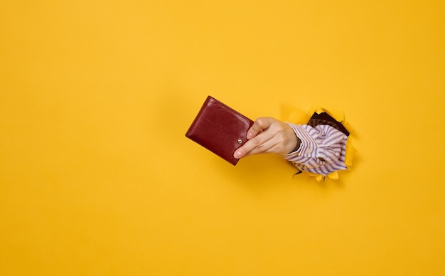 Hand holds a brown leather wallet on a yellow background, part of the body sticks out of a torn hole in the paper