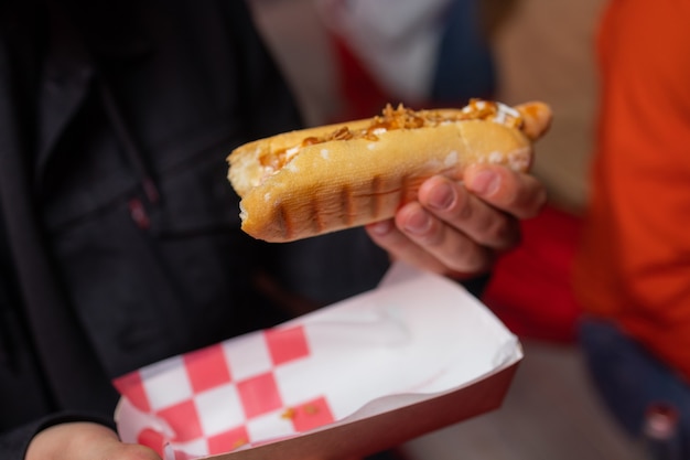 Hand holds fried sausage in dough with ketchup in paper red white packaging on the street