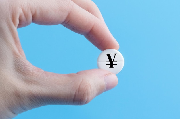 A hand holds a medical pill with an yen or yuan currency sign on a blue background The concept of a rise in the price of medicines in Japan or China