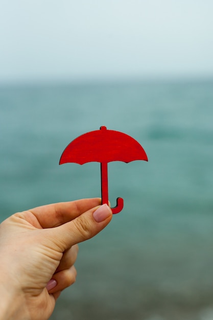 The hand holds a miniature red umbrella against the background of the sea symbol the figure of an um...