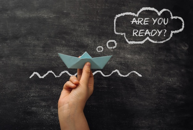 Hand holds paper ship near text are you ready on chalkboard