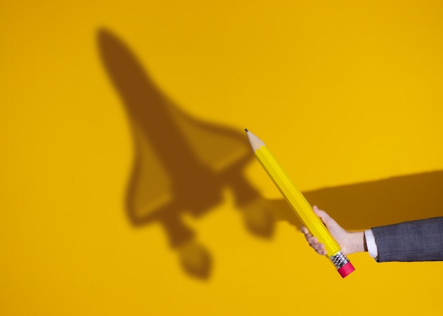 Hand holds a pencil. Shadow of rocket on yellow background. Business startup concept