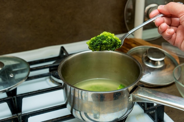 Hand holds a spoon with broccoli over a saucepan on the stove