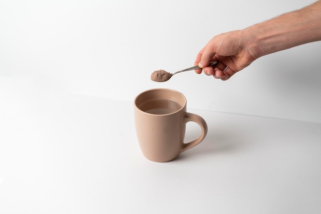 A hand holds a spoon with cocoa powder and put it in cup with hot water