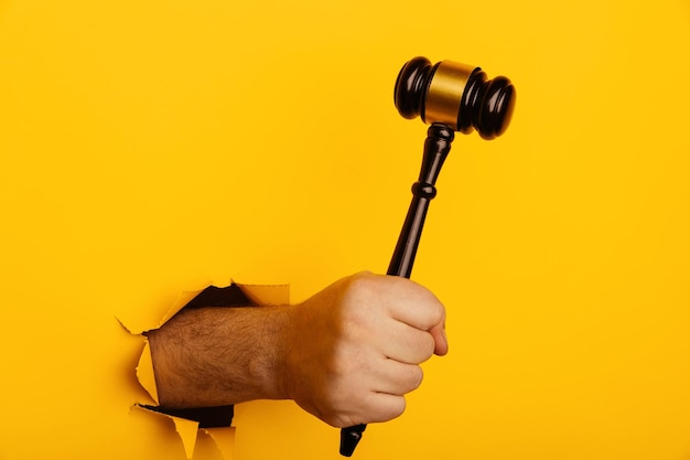 Hand holds a wooden gavel through torn yellow background Law or auction concept