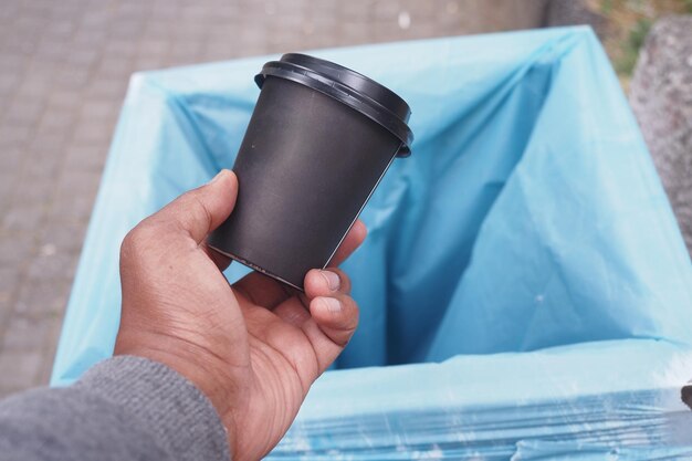 A hand is holding a black coffee cup near a blue waste bin prepared for disposal