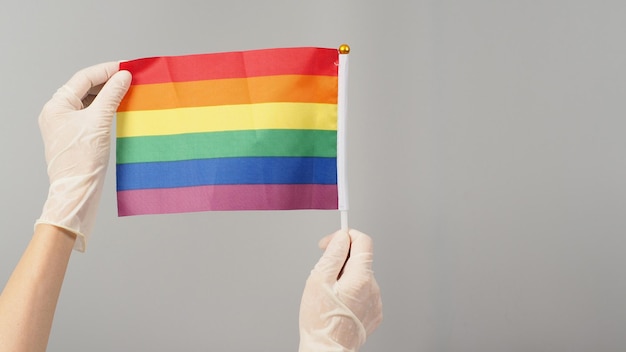 Hand is holding a rainbow flag and wear medical glove on grey background.