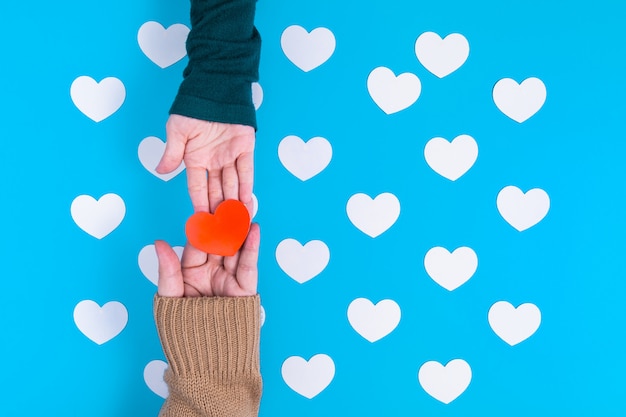 Photo hand is holding on a red heart to the someone hand, those are over a group of white hearts placed on blue