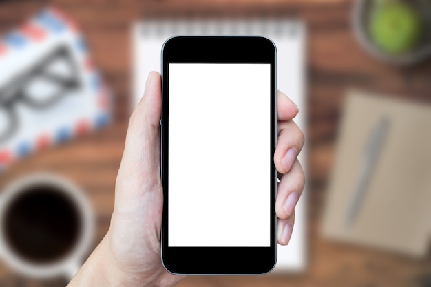 Hand is holding smartphone with blank mockup screen over the office desk.