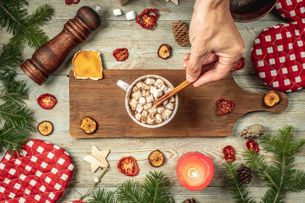 Hand is stirring hot chocolate decorated with marshmallows with a cinnamon stick. Concept of cozy New Year and Christmas mood. Top view