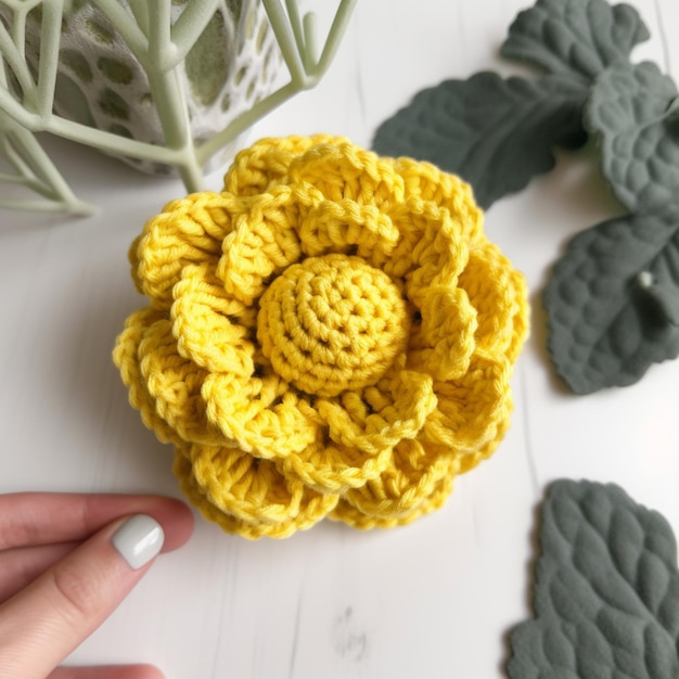 A hand is touching a yellow crochet flower with a green leaf on the side.