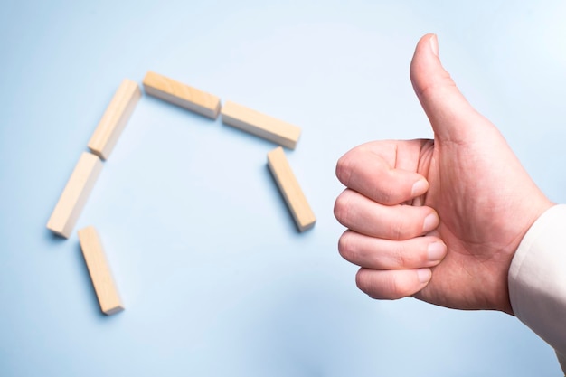 Hand making a thumbs up sign holding his hand inside a house made of wooden pegs. Over yellow background.