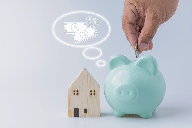 The hand of a man holding putting coins into a green piggy bank for saving money use investment property with a little wood house on white background with copy space Save money financial concepts