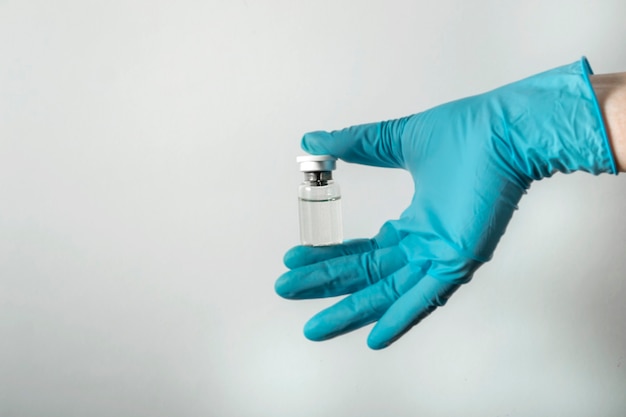 A hand in a medical blue glove holds an ampoule with a vaccine against coronavirus infection COVID-19 on a white background .. Ampoule with a medicine. The doctor holds a vaccine in his hand.