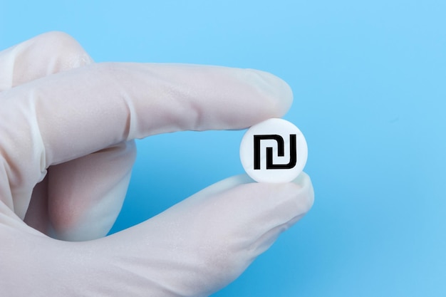 A hand in a medical glove holds a pill with a shekel sign on a blue background Expensive medicine in Israel