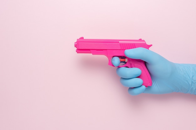 Hand in a medical glove with a pink handgun on pink background