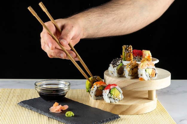 Hand of person holding chopsticks to pick up a sushi roll from a wooden tray with sauce and wasabi