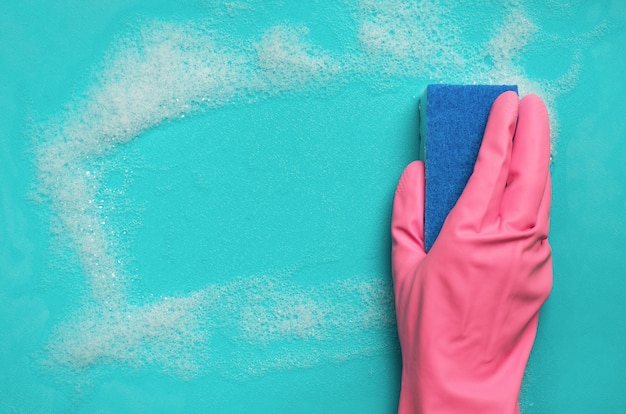 Hand in pink rubber glove holding sponge and wipes soapy foam on blue surface. Cleaning concept