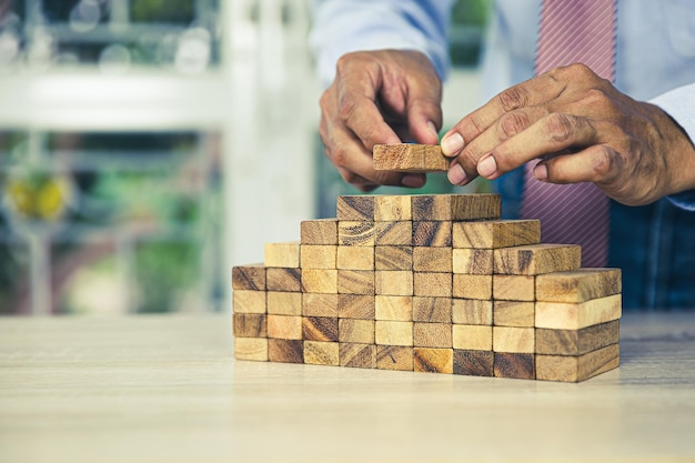 Hand placing wooden block tower stack in pyramid stair step with caution to prevent collapse or crash concepts of financial risk management and strategic planning and business challenge plan