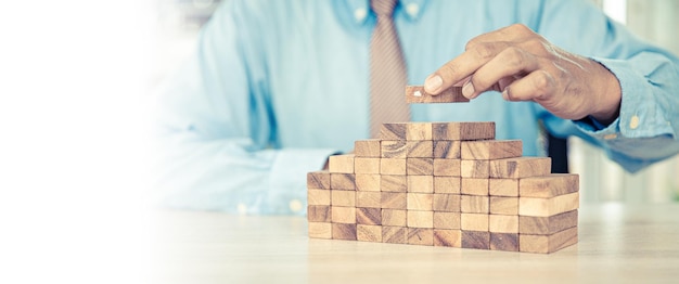 Hand placing wooden block tower stack in pyramid stair step with caution to prevent collapse or crash concepts of financial risk management and strategic planning and business challenge plan