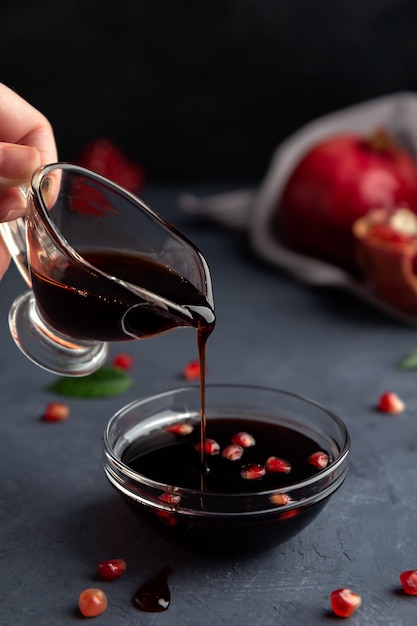 A hand pours pomegranate sauce from a gravy boat into a transparent bowl on dark