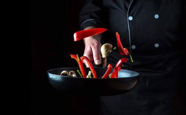 hand of professional chef tosses pieces of vegetables and mushrooms on frying pan