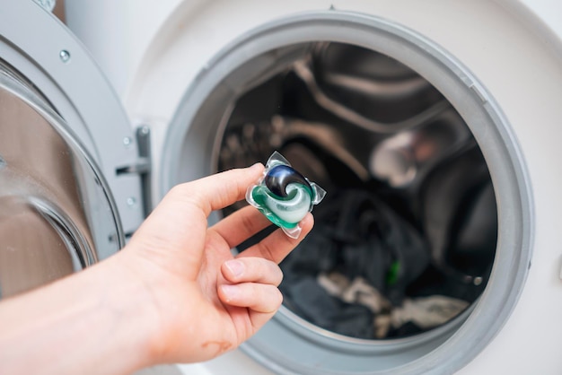 A hand puts laundry powder capsule in washing machine with colorful clothes