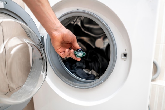 A hand puts laundry powder capsule in washing machine with colorful clothes