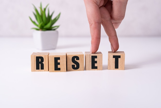 The hand puts a wooden cube with the letter R from the word Reset. The word is written on wooden cubes standing on the yellow surface of the table.