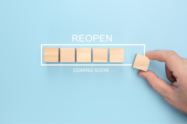 Hand putting wooden cube on virtual infographic loading bar with REOPEN coming soon wording.