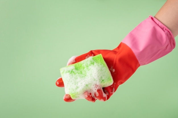 Hand in rubber glove holding green sponge for washing dishes with foam. House cleanining service concept.