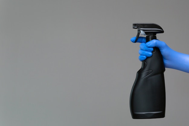 A hand in a rubber glove holds the glass cleaner in a spray bottle on a neutral background.