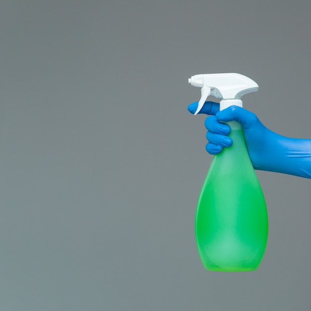 A hand in a rubber glove holds the glass cleaner in a spray bottle on a neutral background. 