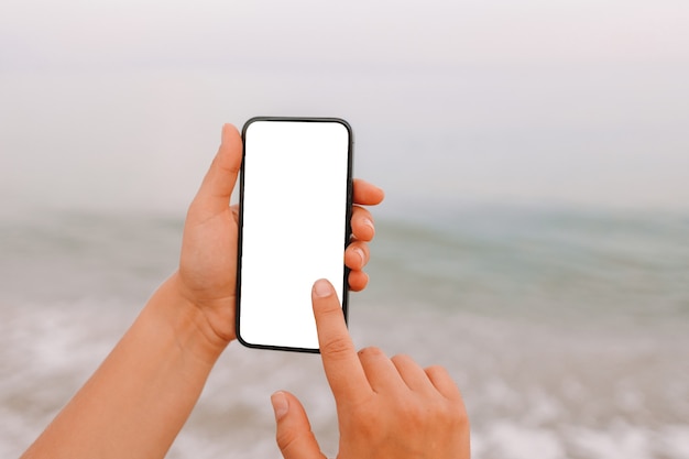 Hand showing a blank smart phone on the beach with the sea in the background. White screen mock up