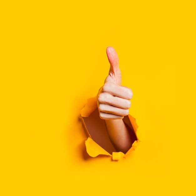 Hand showing a thumb up sign through a ripped hole in yellow paper background Well done good job concept Like gesture