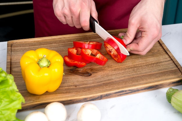 Hand slice red pepper with ceramic knife on wooden cutting board. Vegetarian, health, diet concept. Food preparation, cooking, culinary, cuisine.