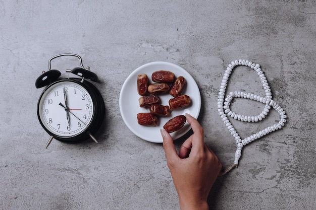 Hand take date fruit arranged with alarm clock showing iftar time and prayer beads.