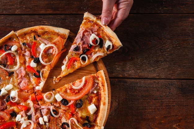 Hand taking hot slice of colorful delicious italian pizza served on wooden table, flat lay. Dark background with copy space.