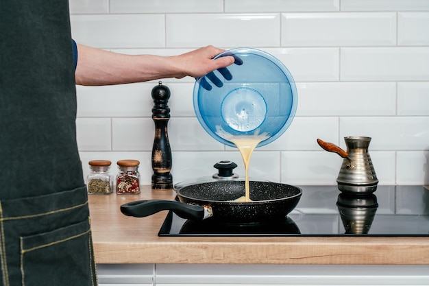 Hand of unrecognizable man in black apron pouring omelette mix from blue plastic bowl in hot frying pan on electric cooker near glass jars with spices spice grinder glass lid and cezve Cooking