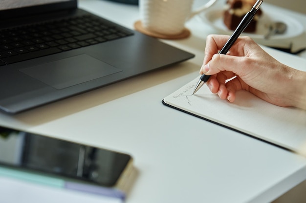 Hand of unrecognizabled person draws sketch on paper sits at home at his desk with laptop in front o