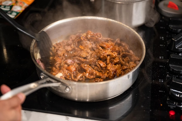 Hand using kitchen flipper frying sliced beef in iron pan on electric stove