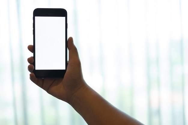 Hand using a mobile phone with white blank screen against white curtain background