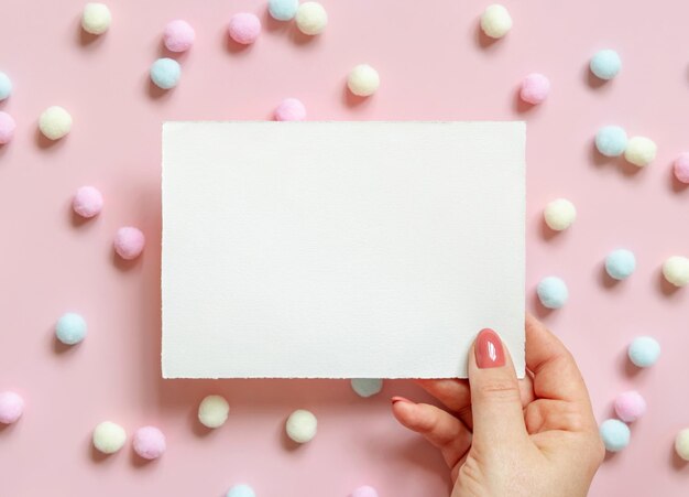 Hand with blank card near pastel pom-poms on pink top view. Romantic scene with blank horizontal card mockup flat lay, place for text . Valentines, Spring or girlish concept