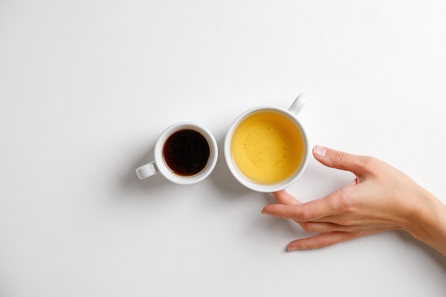 Hand with coffee opposite tea. Mug of black coffee opposite mug of green tea on a white table with hand. Minimalism.